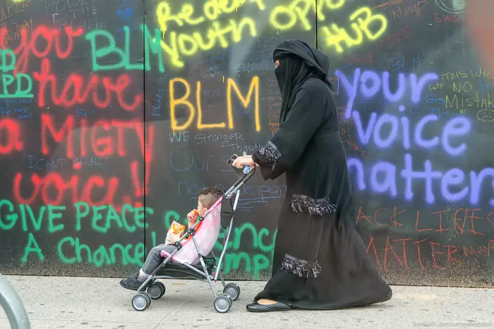 A photo of a woman pushing her child past a wall filled with BLM graffiti